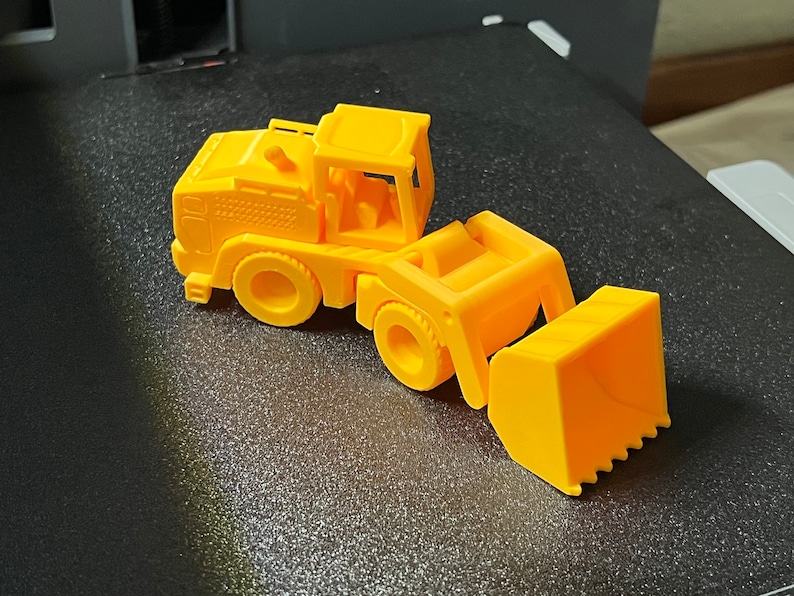 May include: A bright yellow toy front-end loader, featuring a detailed cab, wheels, and a functional-looking bucket. The toy is made of plastic and sits on a dark surface. The design is reminiscent of construction vehicles.