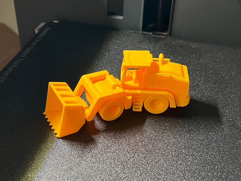 May include: A bright yellow toy bulldozer with a scoop and wheels. The construction vehicle is made of plastic and is sitting on a textured surface. The toy is a miniature replica of a real-life construction machine.