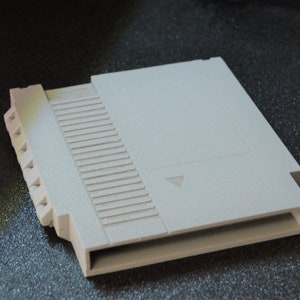 Puede incluir: Un cartucho de plástico blanco para una consola de videojuegos Nintendo Entertainment System (NES). El cartucho tiene una ranura para insertar una tarjeta de juego y un área de etiqueta en la parte superior.