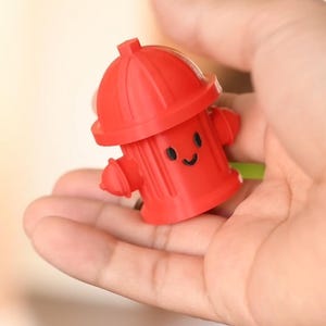 May include: A vibrant red, cartoon fire hydrant toy with a smiling face. The hydrant features a rounded top, two side nozzles, and a small rectangular outlet. The toy is held in a hand.
