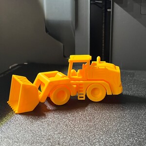 May include: A bright orange toy front-end loader, featuring a scoop, wheels, and a cab. The toy is made of plastic and is designed to resemble a construction vehicle. The toy is sitting on a black surface.