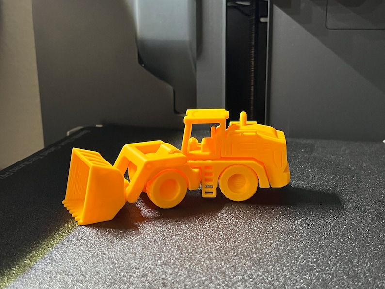 May include: A bright orange toy front-end loader, featuring a scoop, wheels, and a cab. The toy is made of plastic and is designed to resemble a construction vehicle. The toy is sitting on a black surface.