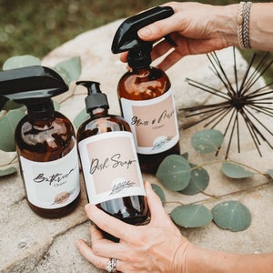 May include: Three amber glass spray bottles with white labels. The labels have handwritten text that reads "Bathroom Cleaner", "Dish Soap", and "Glass & Mirror Cleaner".