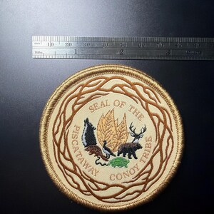 May include: A circular embroidered patch featuring the Seal of the Piscataway Conoy Tribe, with a diameter of approximately 3.5 inches. The design includes a central emblem of leaves, animals, and text, surrounded by a brown vine border.