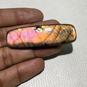 Puede incluir: Una piedra preciosa de labradorita rectangular con una superficie pulida. La piedra muestra un vibrante juego de colores, con tonos rosas, naranjas y dorados. La piedra preciosa se sostiene en una mano, mostrando su tamaño y forma.