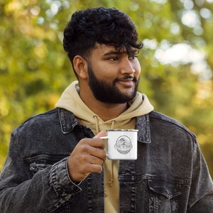 May include: A white enamel camping mug with a black and white graphic of a mountain and trees. The text "SMORE THE MERRIER" is printed on the mug.