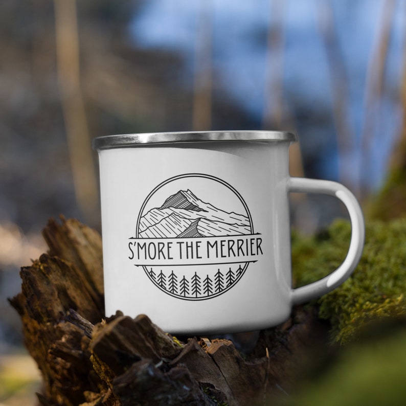 May include: White enamel camping mug with a black and white mountain graphic and the text "S'more the merrier". The mug is sitting on a piece of weathered wood.