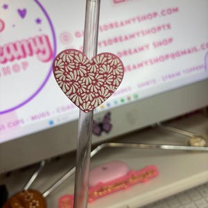 May include: A heart-shaped straw topper in a soft pink hue, adorned with a white floral pattern. The topper is attached to a clear straw, perfect for adding a decorative touch to beverages. The background includes a computer screen with a website address.