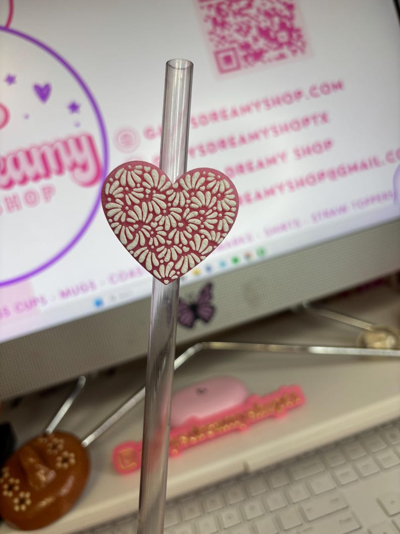 May include: A heart-shaped straw topper in a soft pink hue, adorned with a white floral pattern. The topper is attached to a clear straw, perfect for adding a decorative touch to beverages. The background includes a computer screen with a website address.