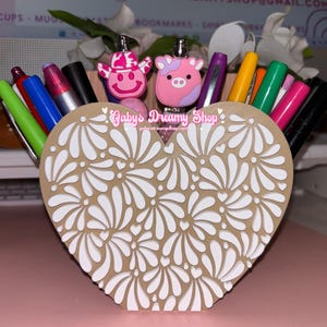 May include: A heart-shaped desk organizer with a floral design, filled with pens and markers. The organizer is tan with white floral cutouts. Two pen toppers shaped like a pink cow and a pink cowboy are visible. The text "Gaby's Dreamy Shop" is printed on the organizer.