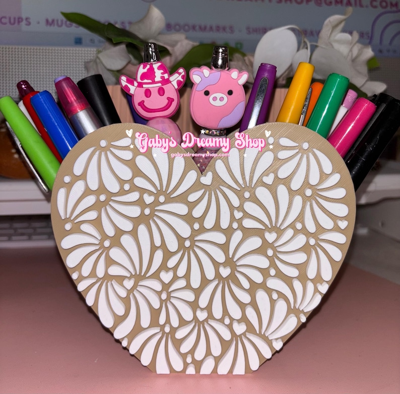 May include: A heart-shaped desk organizer with a floral design, filled with pens and markers. The organizer is tan with white floral cutouts. Two pen toppers shaped like a pink cow and a pink cowboy are visible. The text "Gaby's Dreamy Shop" is printed on the organizer.