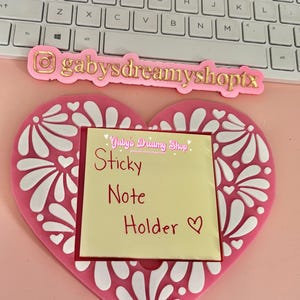 May include: A pink heart-shaped sticky note holder with white floral designs. The holder has a yellow sticky note with the words "Sticky Note Holder" written in red. The holder also has the text "gabysdreamyshoptx" in gold.