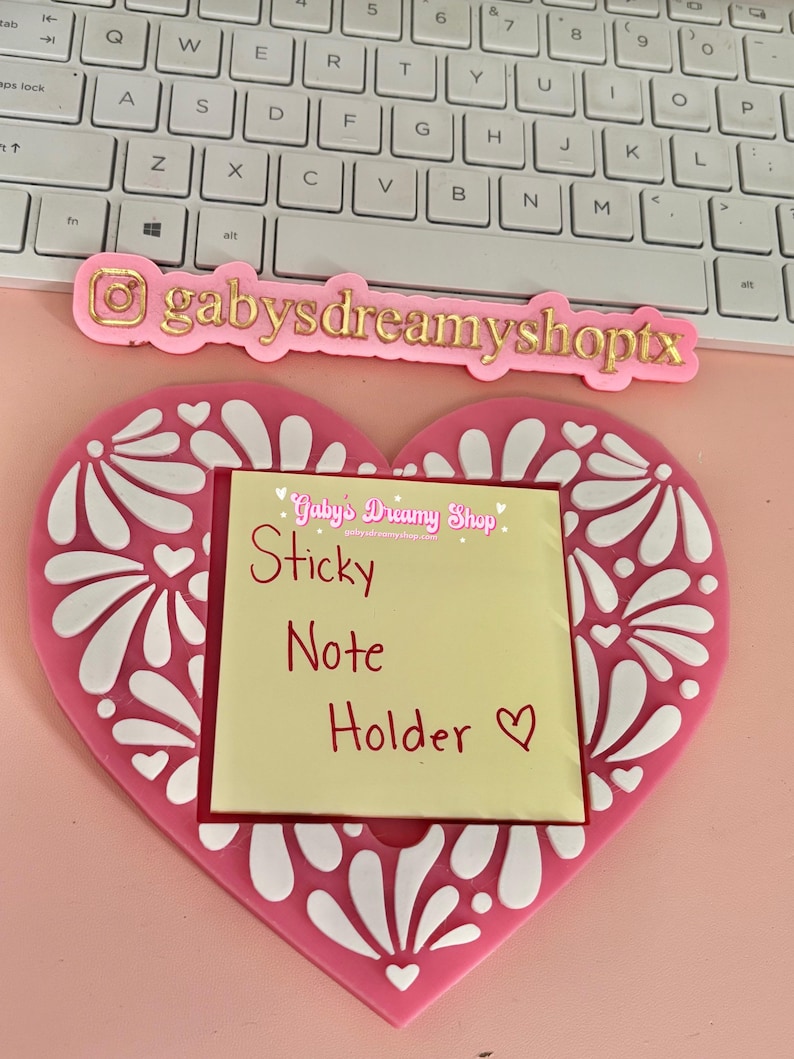 May include: A pink heart-shaped sticky note holder with white floral designs. The holder has a yellow sticky note with the words "Sticky Note Holder" written in red. The holder also has the text "gabysdreamyshoptx" in gold.