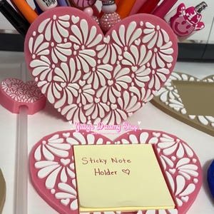 May include: Pink heart-shaped desk organizer with white floral design, holding pens and decorative toppers. A matching sticky note holder with a yellow pad is in front. The text "Sticky Note Holder" is visible.