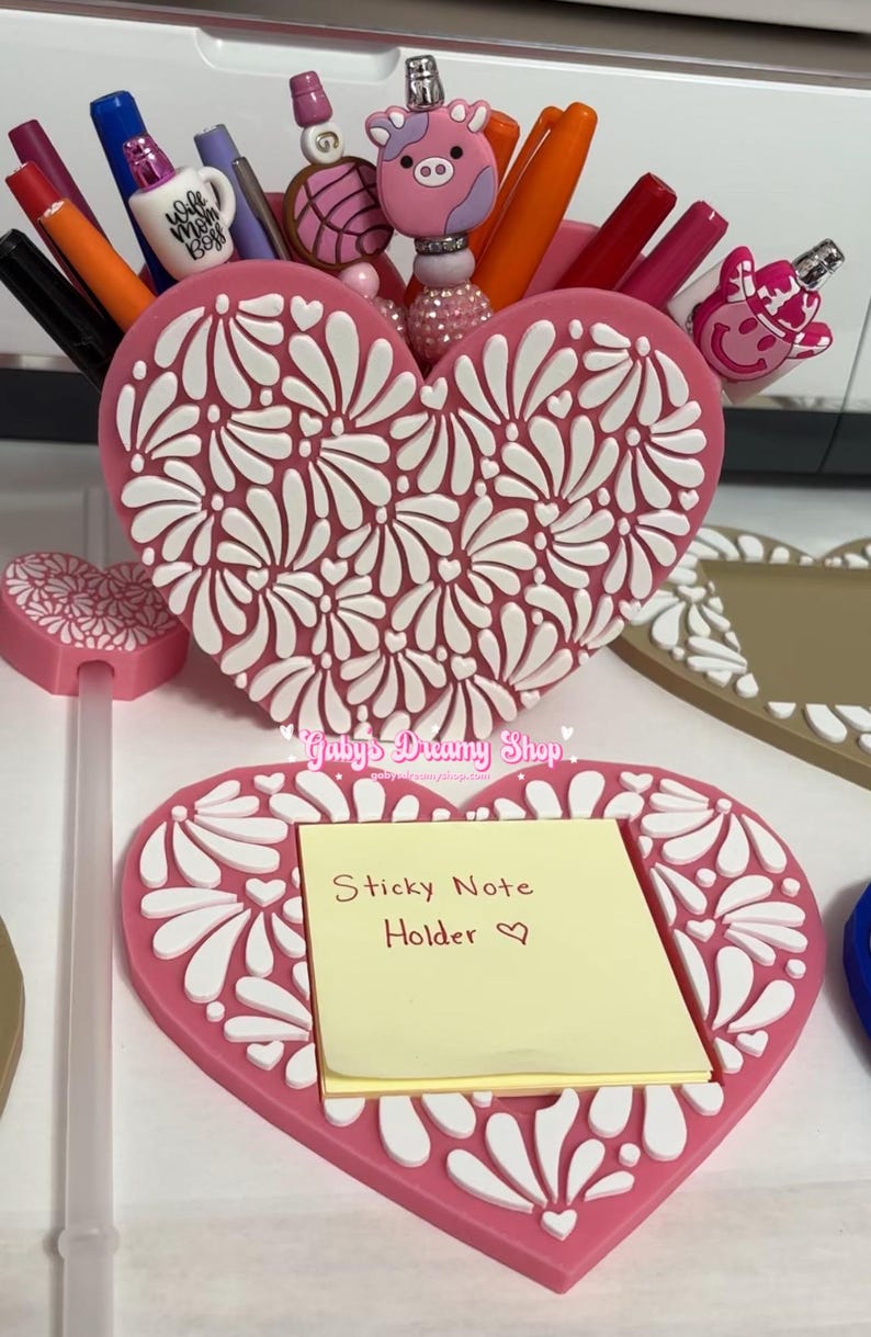 May include: Pink heart-shaped desk organizer with white floral design, holding pens and decorative toppers. A matching sticky note holder with a yellow pad is in front. The text "Sticky Note Holder" is visible.