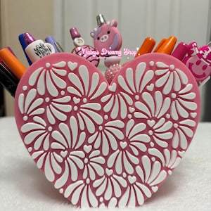 May include: A pink heart-shaped pen holder with a white floral design. The pen holder is filled with pens and markers of various colors. The top of the pen holder has a pink unicorn and other decorative pen toppers. The text "Gaby's Dreamy Shop" is visible.