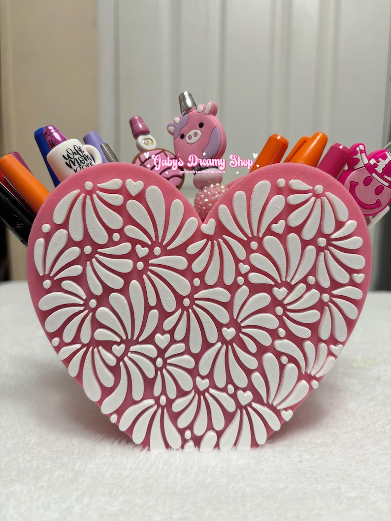 May include: A pink heart-shaped pen holder with a white floral design. The pen holder is filled with pens and markers of various colors. The top of the pen holder has a pink unicorn and other decorative pen toppers. The text "Gaby's Dreamy Shop" is visible.