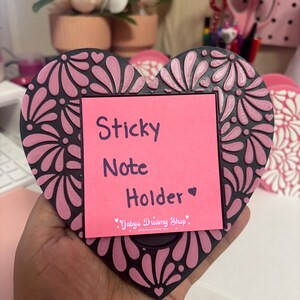 May include: A heart-shaped sticky note holder with a black and pink floral design. The pink sticky note is held in place by a black frame. The text "Sticky Note Holder" is printed on the pink note.