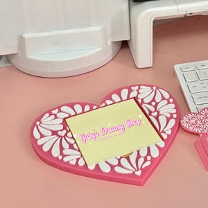 May include: A pink heart-shaped desk organizer with a white floral design. It holds a yellow sticky note pad with the text "Gaby's Dreamy Shop". A keyboard and printer are in the background.