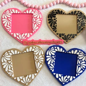 May include: Four heart-shaped frames in pink, tan, and blue, each with a square opening. The frames feature a white floral design around the edges. A pink beaded garland and a small ceramic pot are also visible.