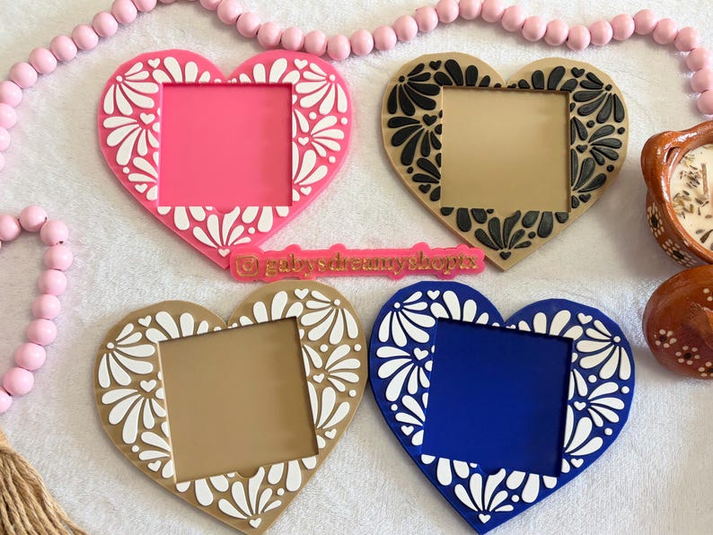 May include: Four heart-shaped frames in pink, tan, and blue, each with a square opening. The frames feature a white floral design around the edges. A pink beaded garland and a small ceramic pot are also visible.