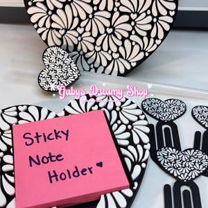 May include: Black and white heart-shaped desk accessories with a floral design. Includes a sticky note holder with a pink pad, a pen holder, and paper clips. The sticky note holder reads "Sticky Note Holder".