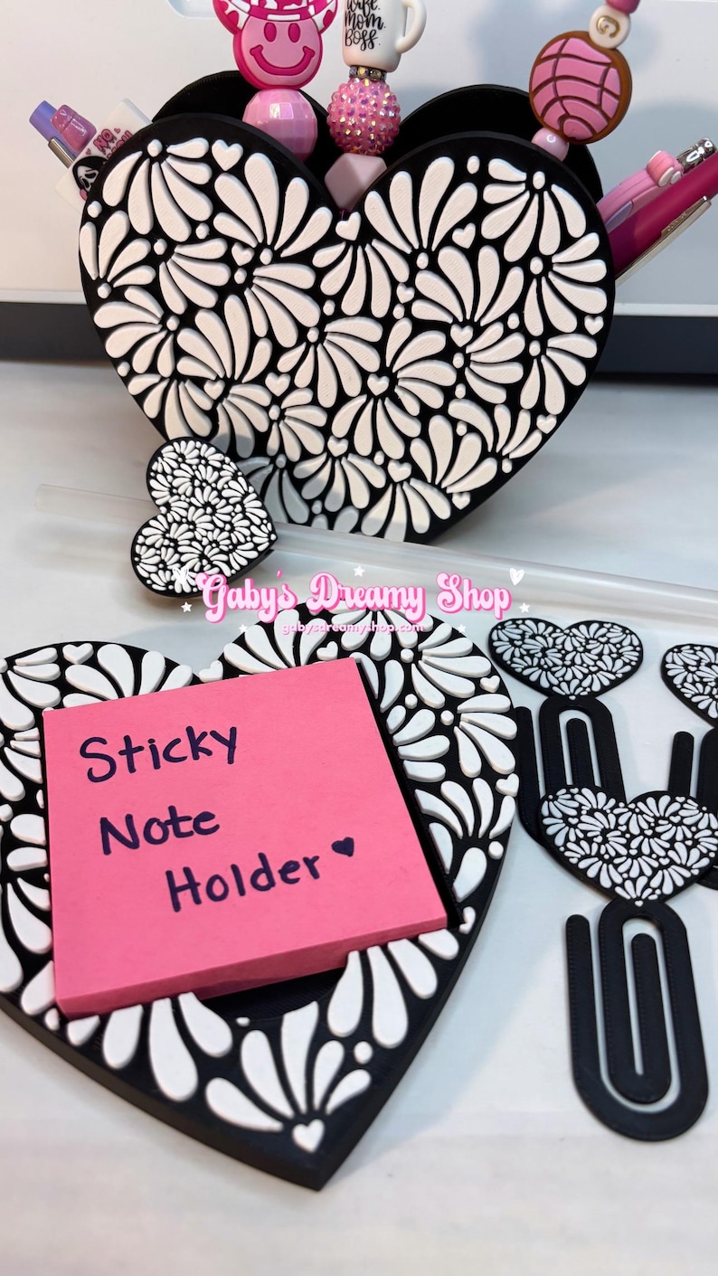 May include: Black and white heart-shaped desk accessories with a floral design. Includes a sticky note holder with a pink pad, a pen holder, and paper clips. The sticky note holder reads "Sticky Note Holder".