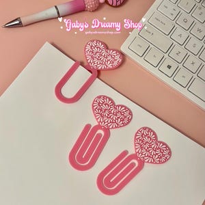 May include: A pink and white pen with a beaded accent and a concha-shaped charm, alongside pink heart-shaped paper clips with white floral designs. The text "Gaby's Dreamy Shop" and website address are visible.