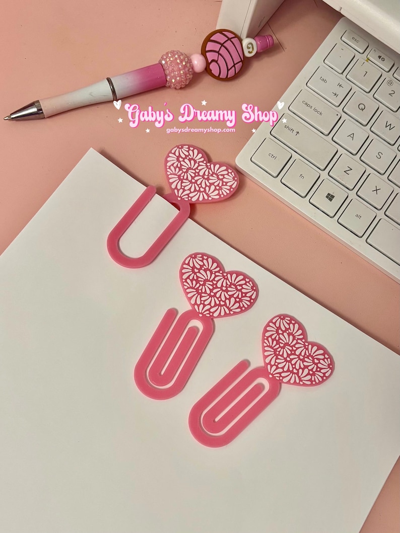 May include: A pink and white pen with a beaded accent and a concha-shaped charm, alongside pink heart-shaped paper clips with white floral designs. The text "Gaby's Dreamy Shop" and website address are visible.