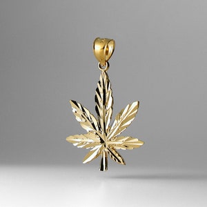 Puede incluir: Un colgante de color dorado en forma de hoja de marihuana. La hoja está detallada con un diseño texturizado.