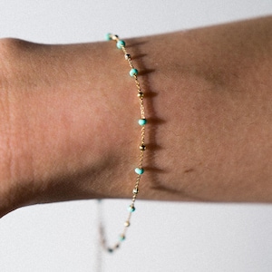 Può includere: Un delicato braccialetto con catena dorata ornato da piccole perle turchesi e dettagli dorati. Il braccialetto è mostrato su uno sfondo neutro, evidenziando la sua semplice eleganza. Un gioiello raffinato.