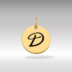 Pode incluir: Pingente circular dourado com uma argola no topo. O pingente apresenta uma letra "D" preta em itálico no centro. O fundo é um gradiente de cinza claro a escuro.