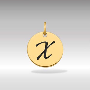 Puede incluir: Un colgante redondo de color dorado con la letra cursiva "X" en negro. El colgante tiene un pequeño lazo en la parte superior para una cadena. El fondo es un degradado de gris claro a blanco.
