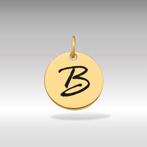 Puede incluir: Colgante circular dorado con un lazo en la parte superior. El colgante presenta una letra "B" negra en cursiva en el centro. El fondo es un degradado de gris claro a oscuro.