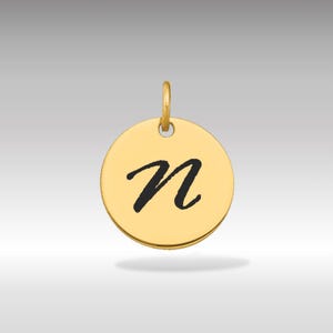 Pode incluir: Pingente circular dourado com a letra "n" preta em itálico. O pingente tem uma pequena argola na parte superior para prender a uma corrente. O fundo é um gradiente cinza claro.