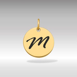 Pode incluir: Pingente circular dourado com a letra "M" preta em itálico. O pingente tem uma pequena argola na parte superior para uma corrente. O fundo é um gradiente cinza claro.