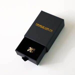 May include: A gold ring with the word "You" written in cursive script, inside a black jewelry box with the words "Charlie and Co. Jewelry" printed in gold.