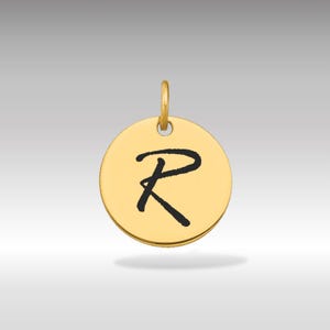 Puede incluir: Un colgante circular de color dorado con un lazo en la parte superior. El colgante presenta la letra "R" negra en cursiva en el centro. El fondo es un degradado de gris claro a gris más oscuro.