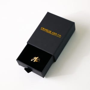 Op de afbeelding: Een zwarte ringdoos met de tekst "CHARLIE AND CO. JEWELRY" in goud gedrukt op de bovenkant. De doos is open en een gouden ring is zichtbaar in de doos.
