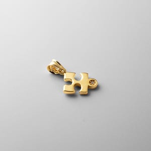Puede incluir: Un colgante de pieza de rompecabezas de tono dorado con un pequeño diamante en el centro.