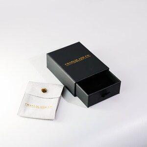 May include: Black jewelry box with a drawer and a white velvet pouch. The box has the text "Charlie and Co. Jewelry" printed in gold on the top.