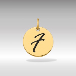 Puede incluir: Un colgante redondo de color dorado con la letra cursiva "Z" negra en el centro. El colgante tiene un pequeño lazo en la parte superior para una cadena. El fondo es un degradado gris claro.