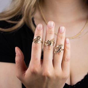 May include: A hand wearing three gold rings with different initial designs. The rings are on the index, middle, and ring fingers.