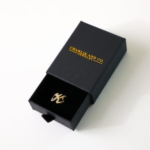 K&ouml;nnte beinhalten: Ein goldener Ring mit einem einzigartigen Design wird in einer schwarzen Schachtel mit dem in Gold gedruckten Text "Charlie and Co. Jewelry" pr&auml;sentiert.