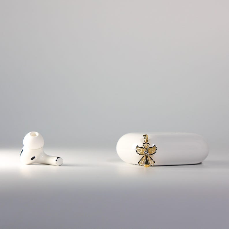 May include: A white wireless earbud charging case with a gold angel pendant on a white surface.