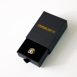 Puede incluir: Un anillo de oro con un dise&ntilde;o delicado e intrincado se muestra en una caja de joyer&iacute;a negra. La caja est&aacute; etiquetada con "Charlie and Co. Jewelry."