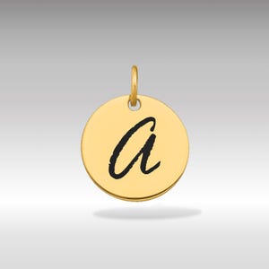 Puede incluir: Un colgante circular dorado con un lazo en la parte superior. El colgante presenta la letra minúscula "a" en negro, en escritura cursiva. El fondo es un degradado de gris claro.