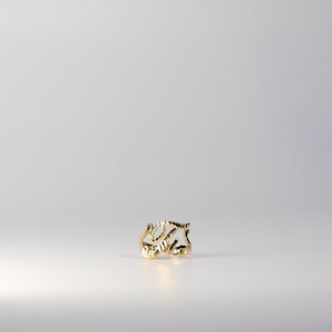 Può includere: Un anello color oro con un design aperto. L'anello presenta una forma stilizzata della lettera "T" con dettagli intricati. L'anello è posto su uno sfondo bianco, evidenziando la sua lucentezza metallica. Gioielli.