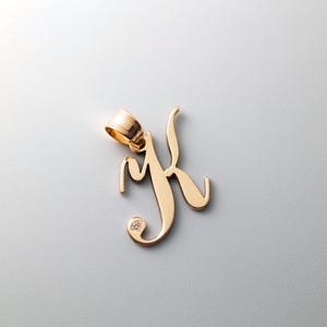 Può includere: Un ciondolo color oro a forma di lettera "K" con un piccolo diamante. Il ciondolo ha un anello nella parte superiore per una catena ed è esposto su uno sfondo grigio chiaro. Il ciondolo è alto circa 2,5 cm.