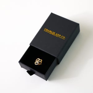 May include: A gold ring with a delicate, intricate design is displayed in a black box with the text "CHARLIE AND CO. JEWELRY" printed in gold.